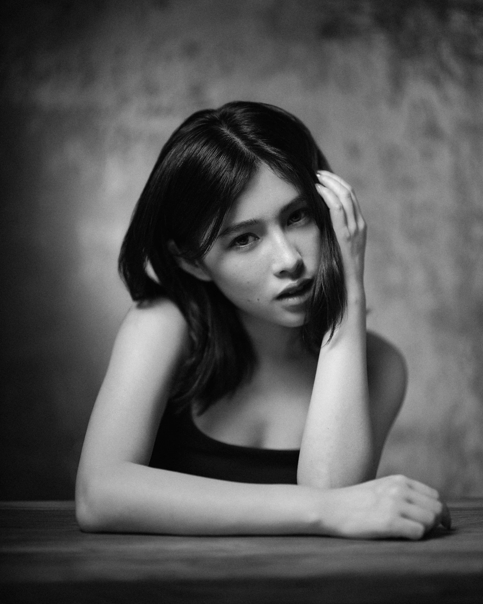 Elegant studio portrait of a woman leaning on a dark surface, wearing a cream textured button-down shirt against a moody, mottled grey background.