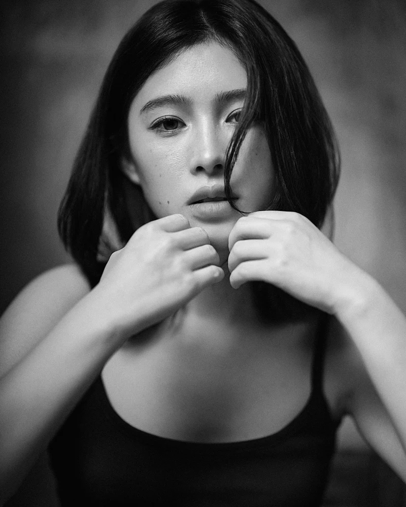 Intimate monochrome studio headshot of a woman resting her chin gently on her clasped hands, featuring soft directional lighting and a dark textured backdrop.