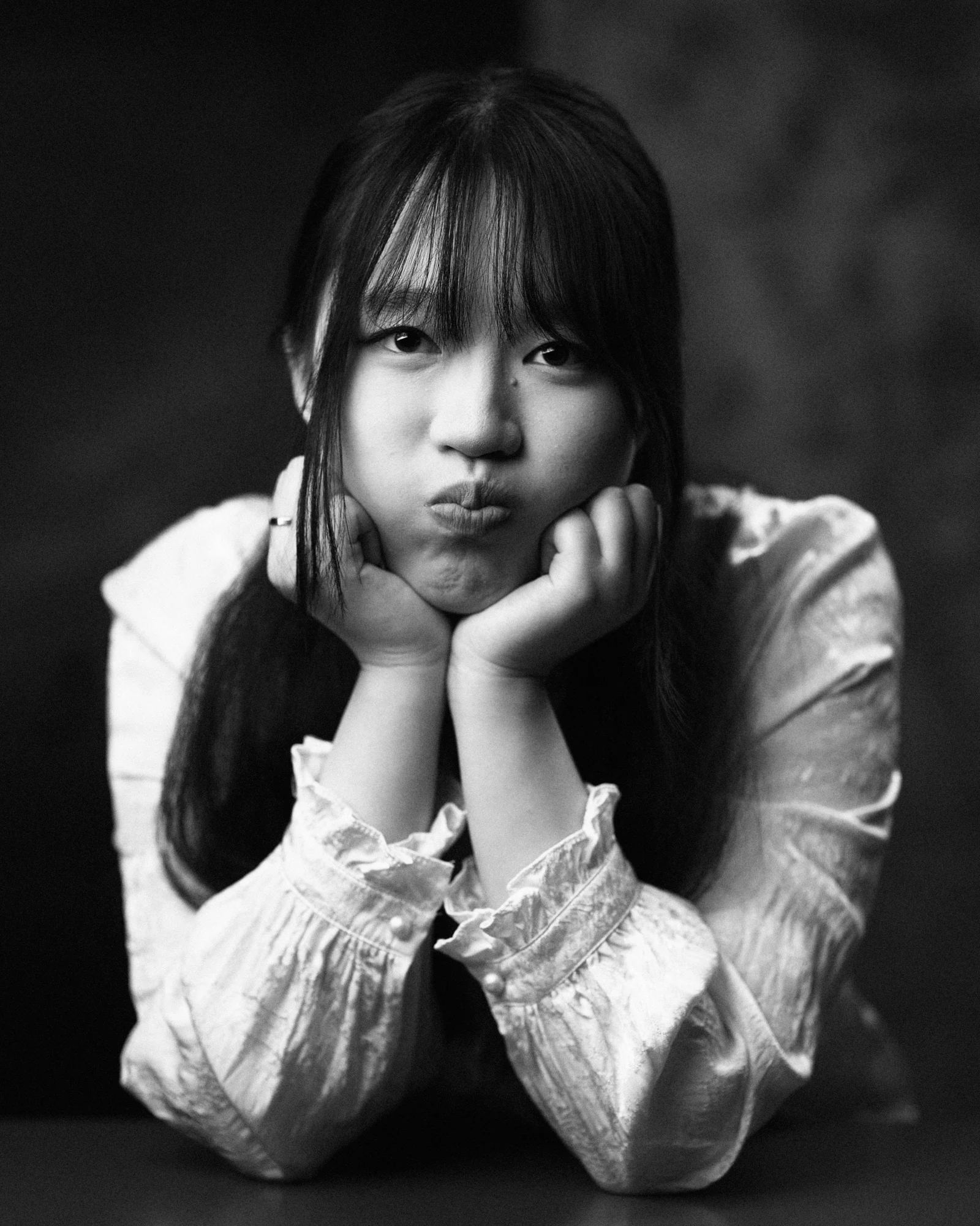 Charming black and white studio portrait of a young woman playfully puffing out her cheeks while resting her chin on both hands, wearing a ruffled white long-sleeve shirt.
