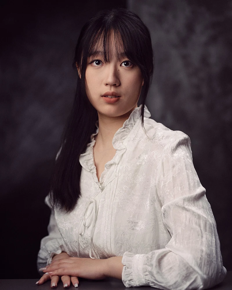 Classic fine art studio portrait of a woman with bangs looking directly at the camera, dressed in an elegant white textured blouse against a moody grey background.