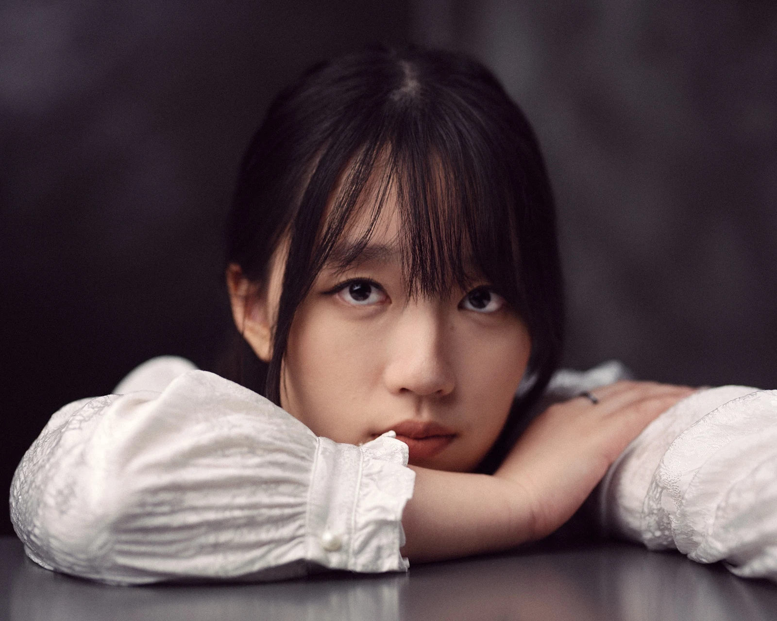 Intimate close-up studio portrait of a young woman resting her chin on her crossed arms on a table, featuring a soft-focus background and soulful expression.