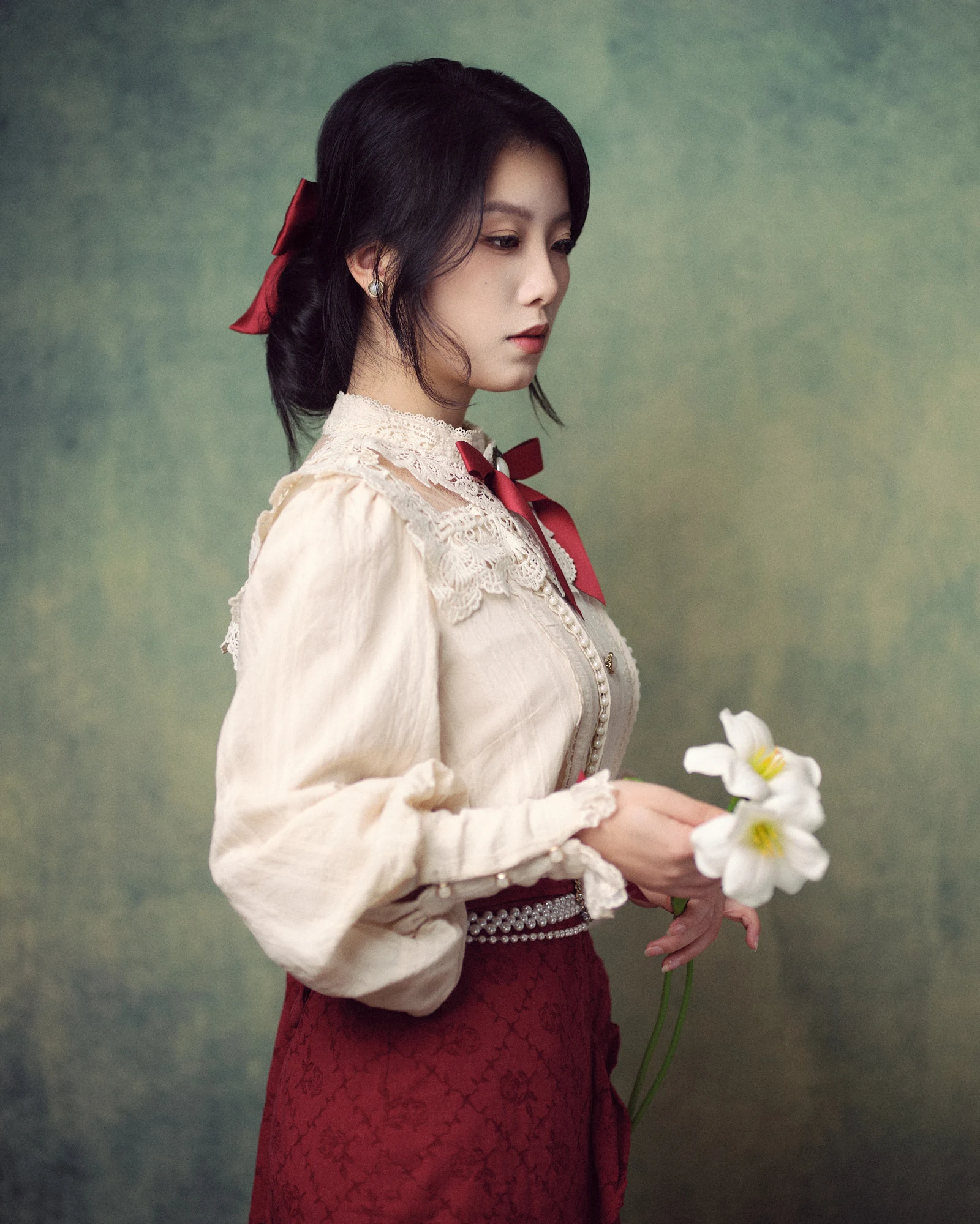Elegant side profile portrait of a young woman dressed in a vintage cream lace blouse and red skirt, holding white lilies in her hands against a textured, olive-green painted backdrop.