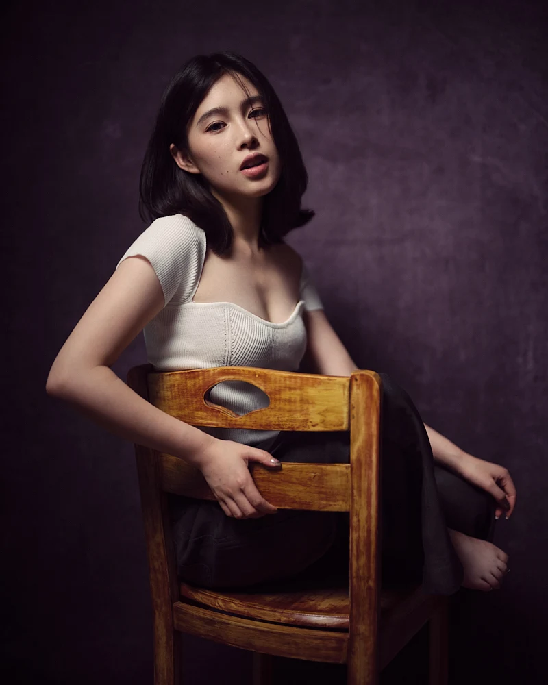 Moody, low-key fine art portrait of a woman with a thoughtful gaze, draped in soft shadows against a deep, dark textured background.